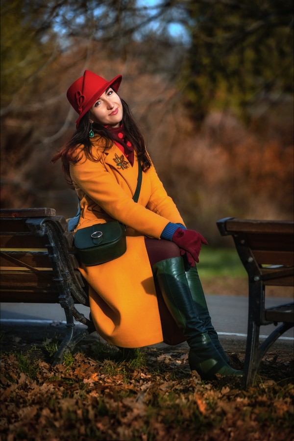 Девушка на лавочке в красной шляпе и желтом пальто улыбается осеннему солнцу