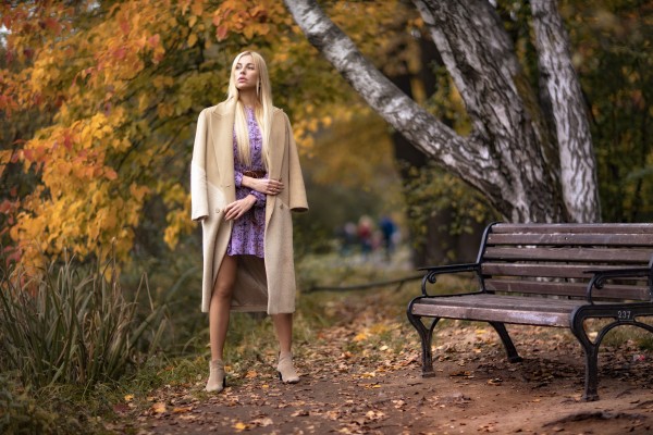 Девушка в бежевом пальто мечтательно смотрит вдаль в осеннем парке