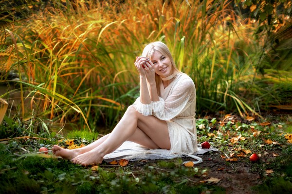 Девушка в белом платье радуется на осенней траве красным яблокам